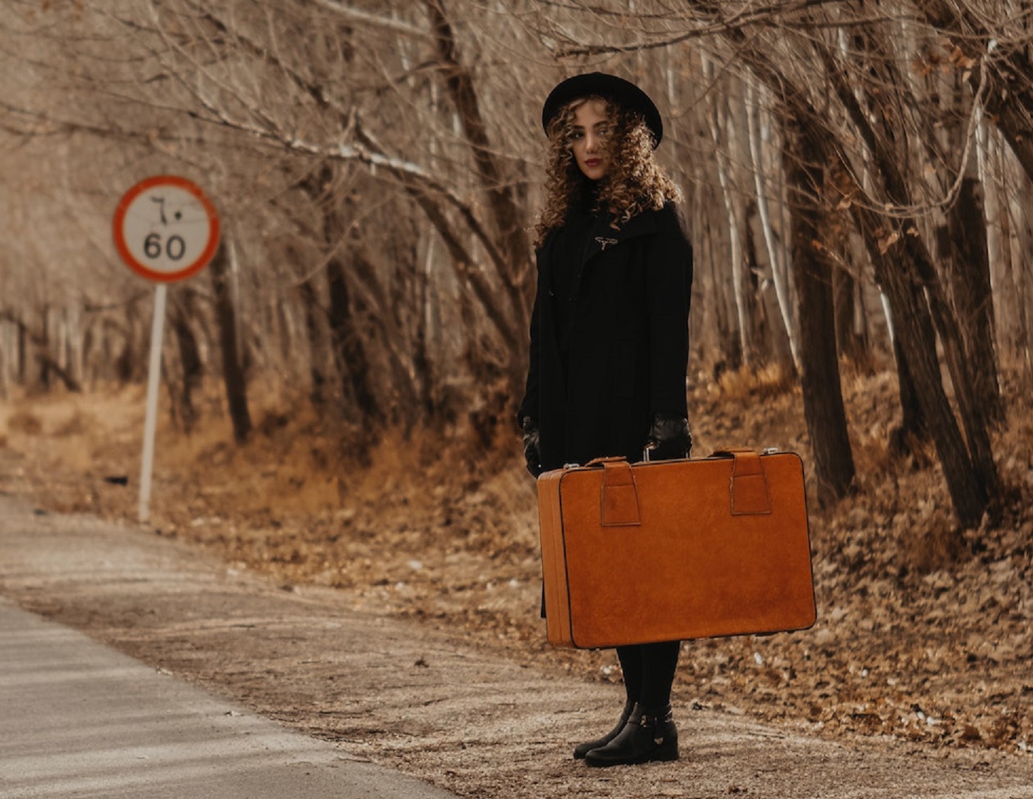 Young woman is holding a brown suitcase, standing by the road.