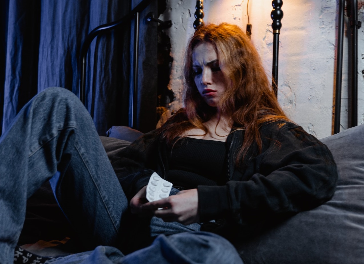 Woman is seating on the bed with pills in her hands, looking upset.