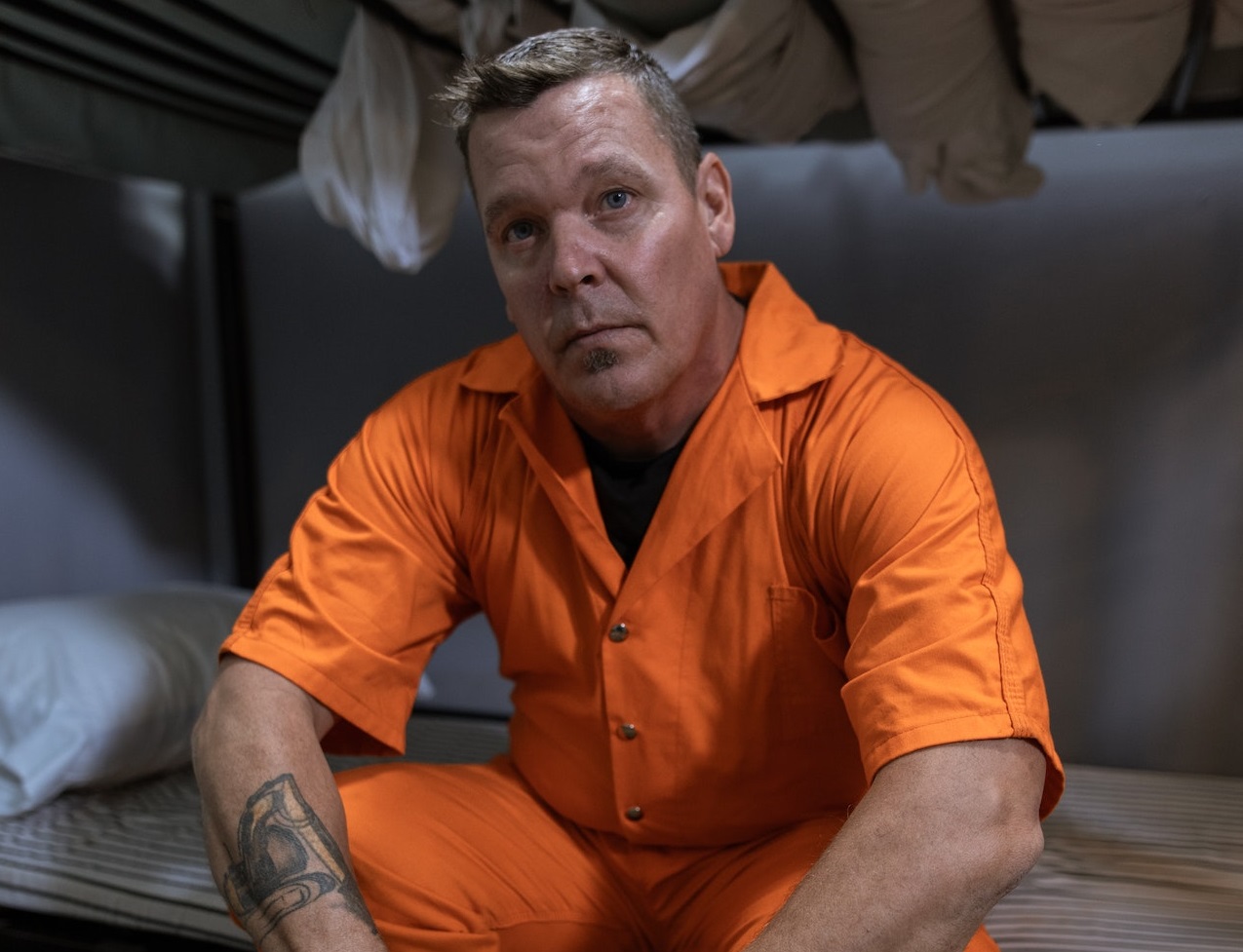 Man is seating in jail cell and looking at camera.