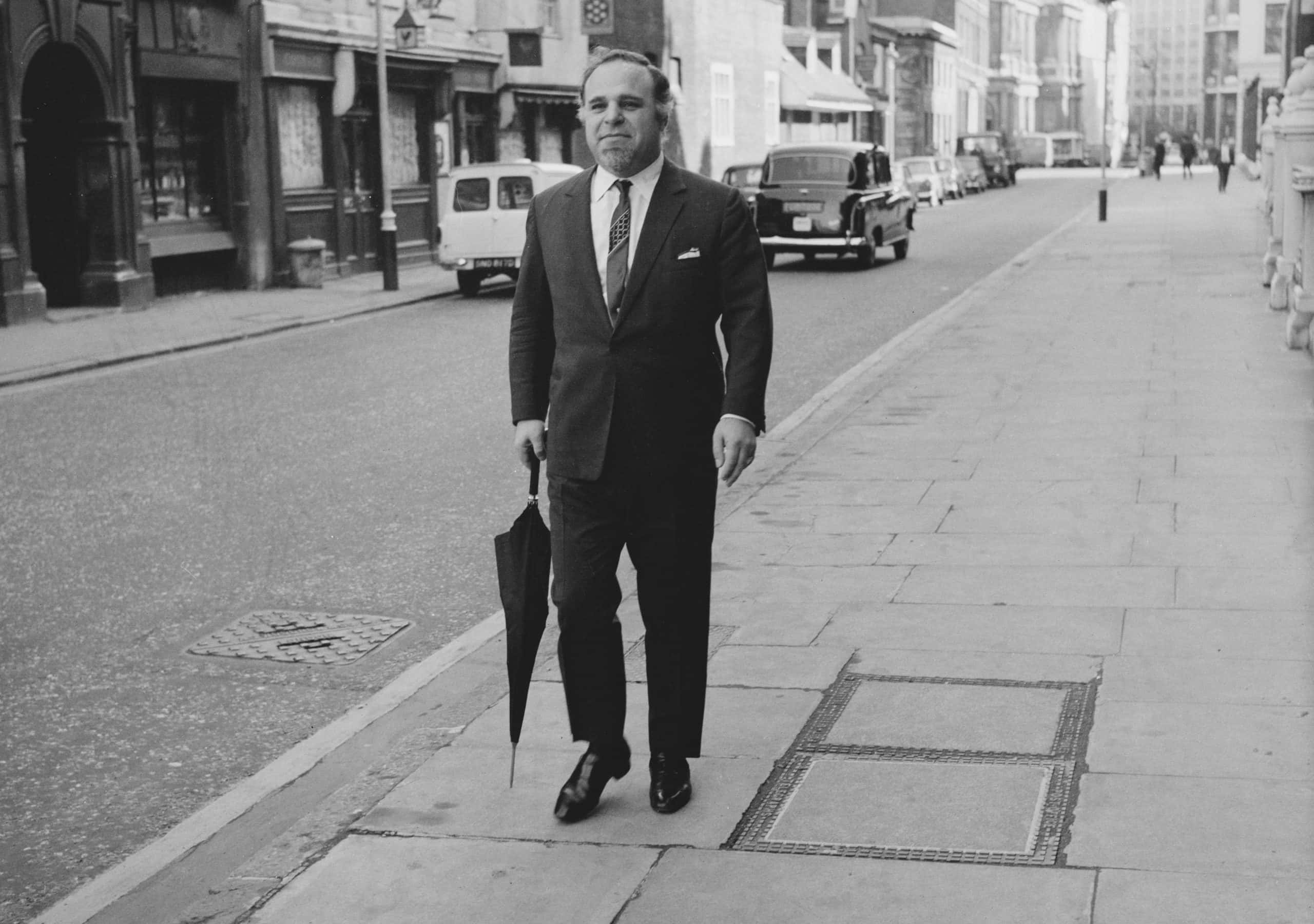 Don Arden in suit and carrying an umbrella walking down the street