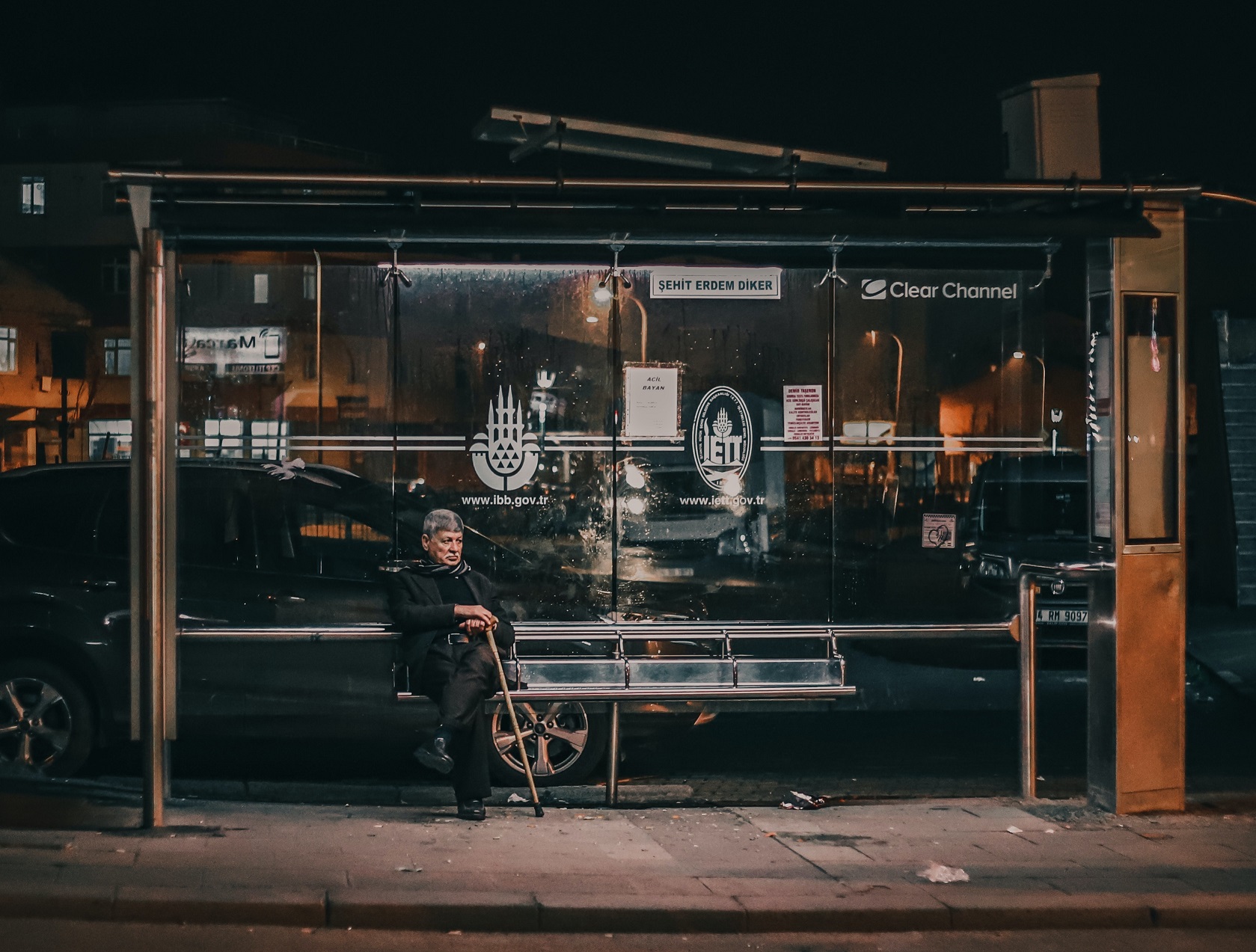 Man wearing black coat is waiting a bus at night, seating on bench.