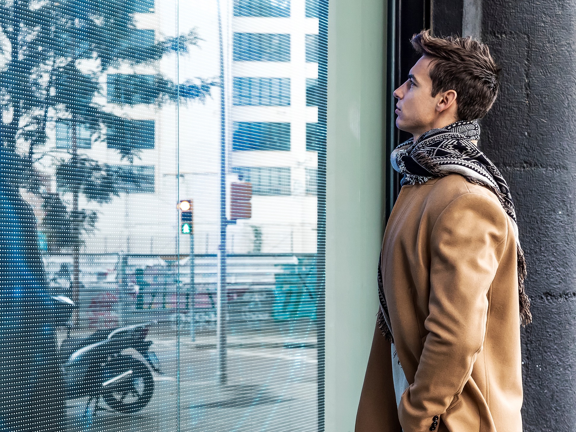 Man wearing brown coat is standing by shop window and looking inside.