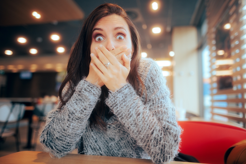Shocked Woman Covering Her Mouth Sitting in a Restaurant in gray sweater