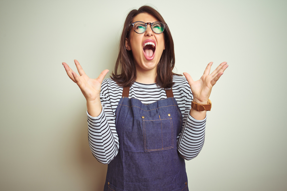 Young beautiful business woman wearing store uniform apron over isolated background crazy and mad shouting and yelling