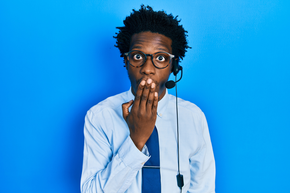 Young African American man wearing call center agent headset covering mouth with hand, shocked in blue shirt