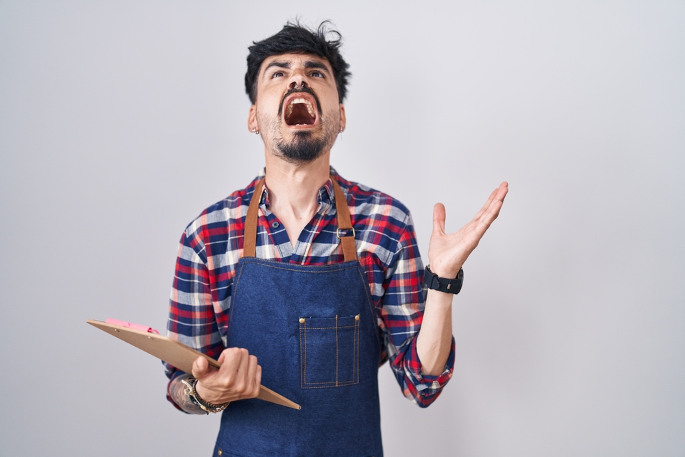 Angry Young Hispanic man with beard wearing waiter apron and plaid shirt holding clipboard, yelling at the camera