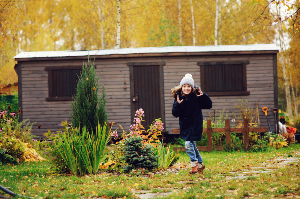 happy kid girl in warm clothes running to wooden shed in garden