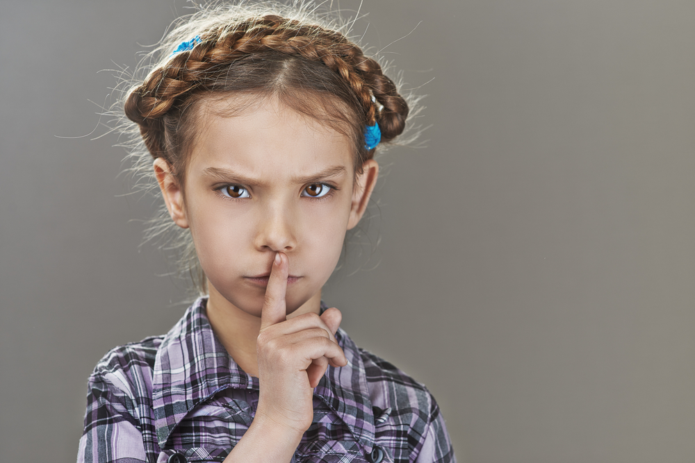 Beautiful sad little girl puts index finger to lips to keep a secret, in plaid shirt , on gray background