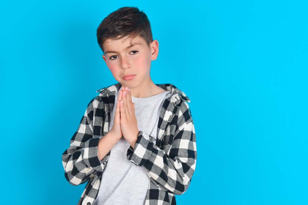 Sad kid  having regretful look, asking for forgiveness in plaid shirt in front of blue background