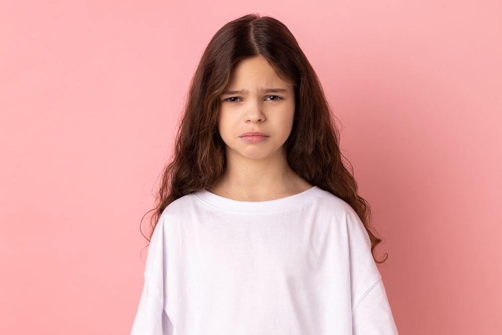 Portrait of upset little girl wearing white T-shirt expressing negative emotions and sorrow in front of pink background