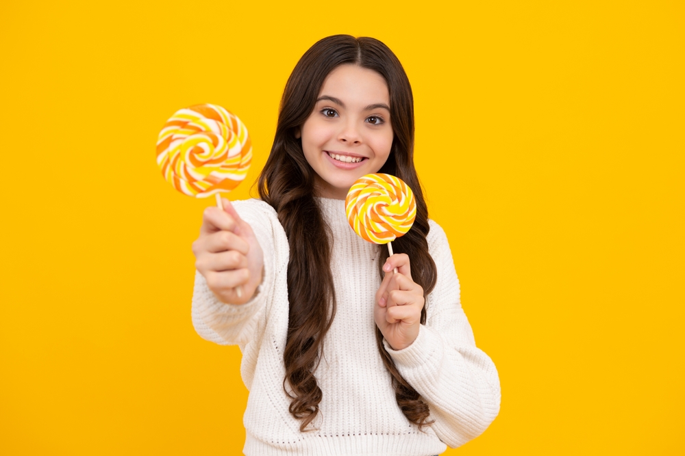 Girl in white sweater giving sweets suckers to her classmates in front of yellow background