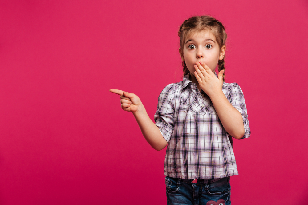 Image of surprised shocked cute little girl in plaid shirt and jeans pointing left with finger