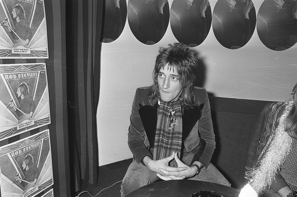 Rod Stewart in jacket and scarf sitting on a couch at  an award ceremony