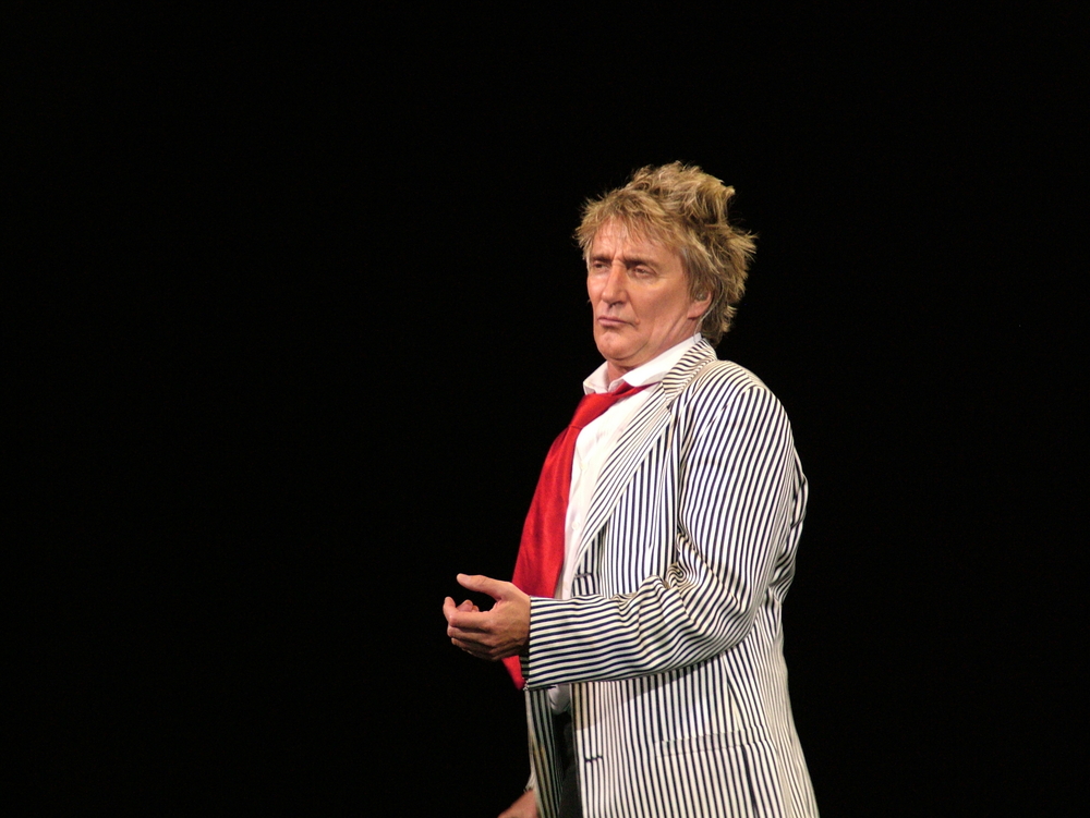 Rod Stewart in striped jacked and red tie performing on stage