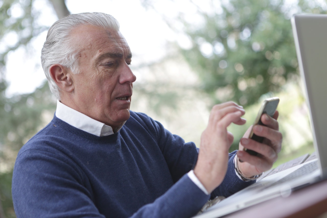 Senior man wearing white shirt and blue sweater is looking at his cell phone.