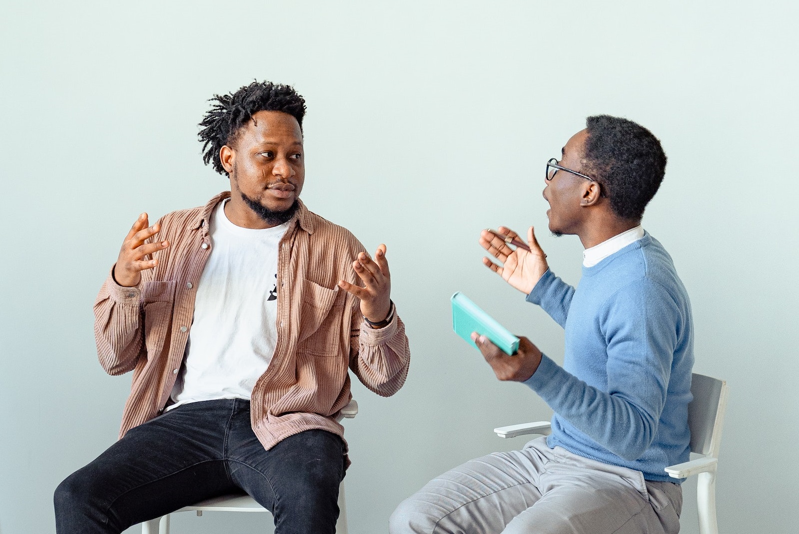 Young black man wearing brown jacket is arguing with other man wearing white shirt and blue sweater, seating on the chairs.