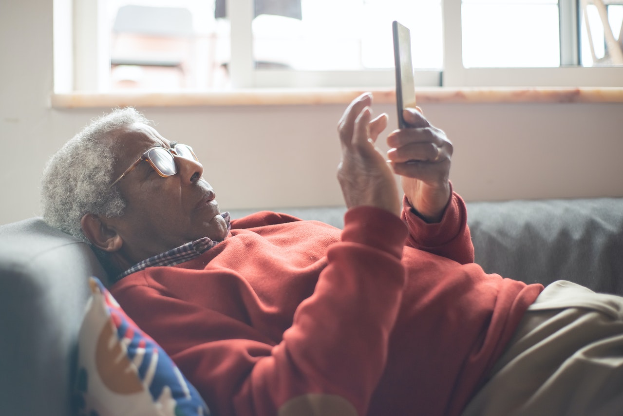 Senior man is laying on the bed and holding a cell phone.
