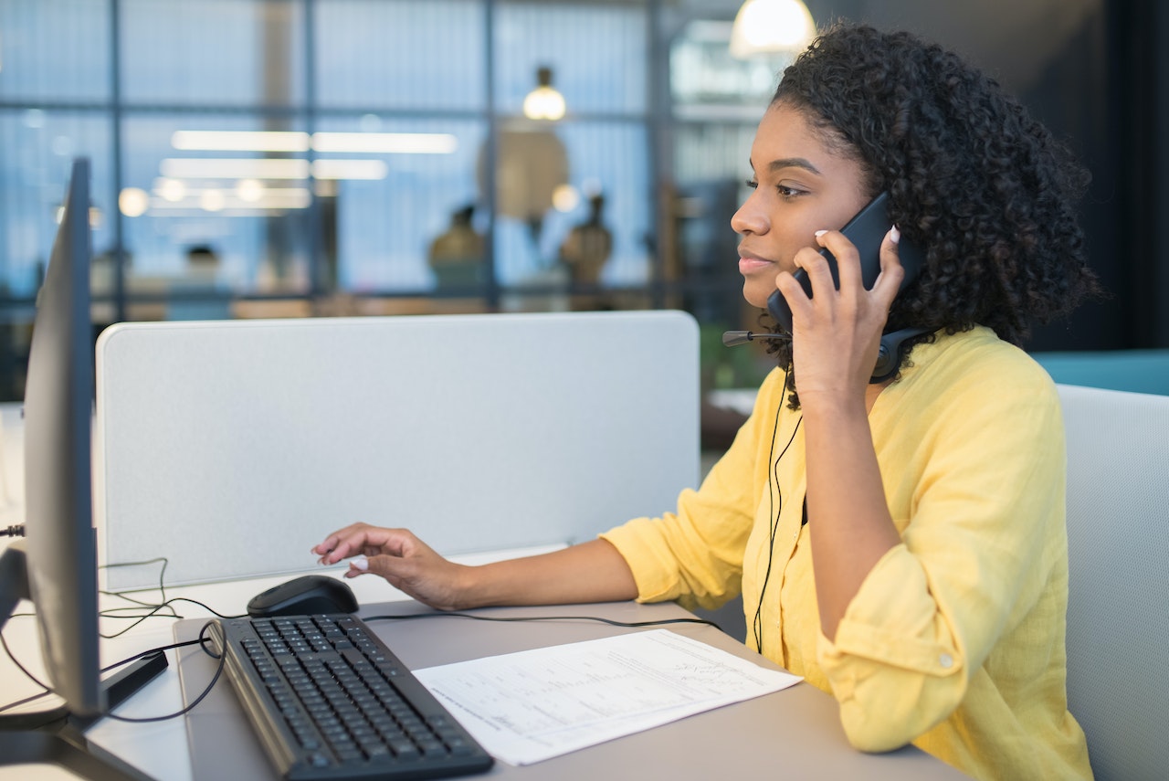 Woman wearing yellow shirt is talking on the phone at her desk.