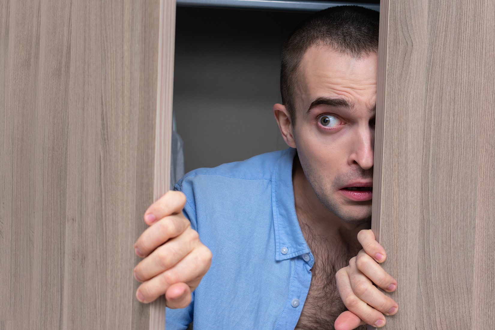 Young man wearing blue shirt is hiding inside a closet.