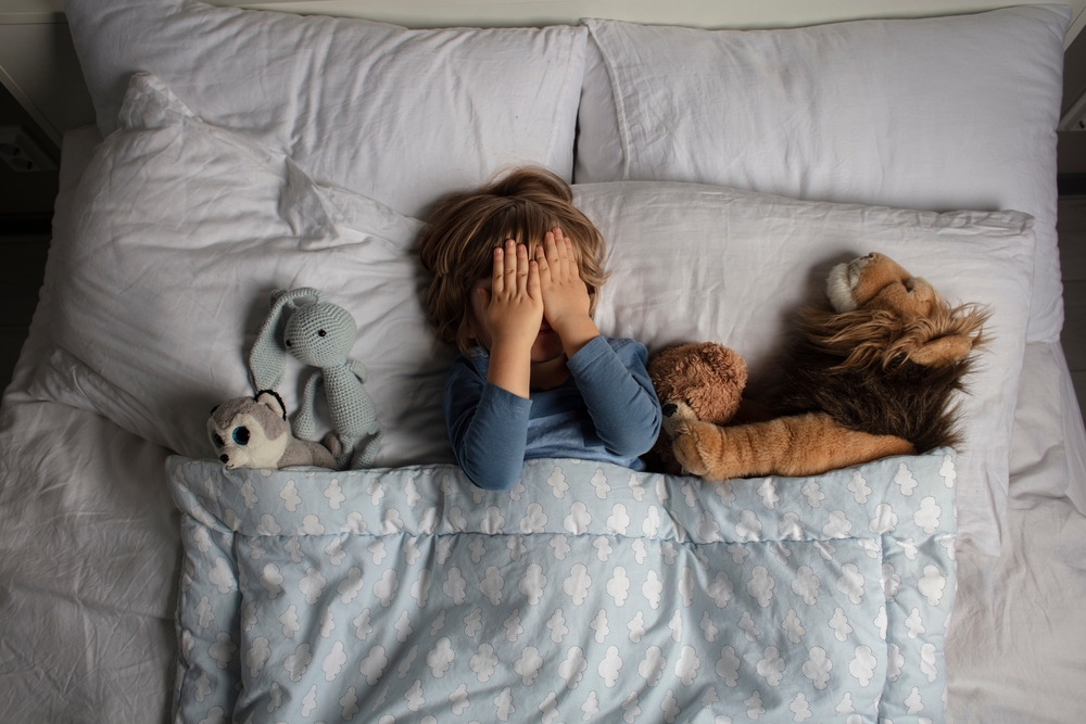 Scared boy covering his face in bed surrounded by toys