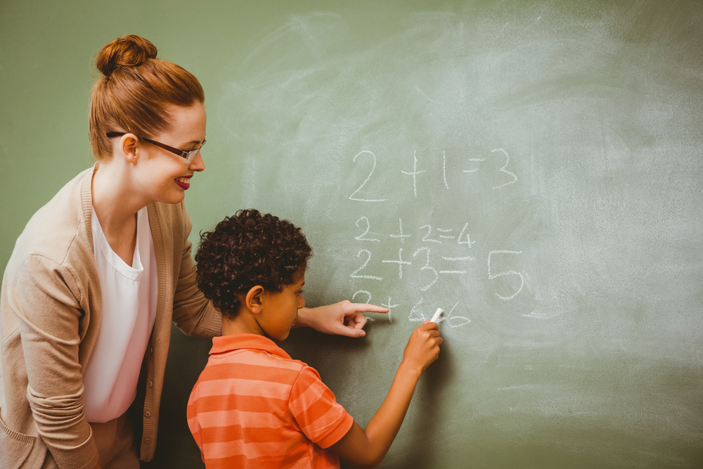 Rear view of teacher assisting little boy to write on blackboard in the classroom