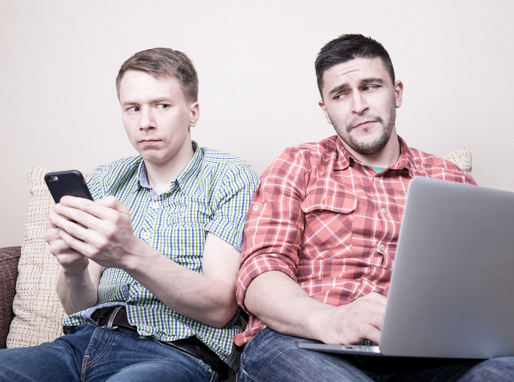 Two young guys operating gadgets sitting on sofa at home