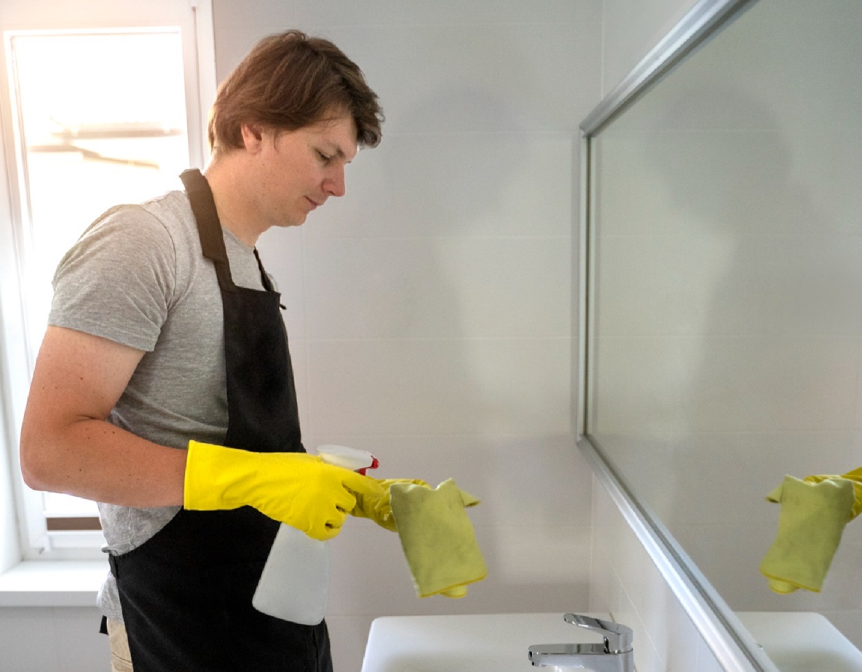 Young man is cleaning the bathroom sink and mirror.