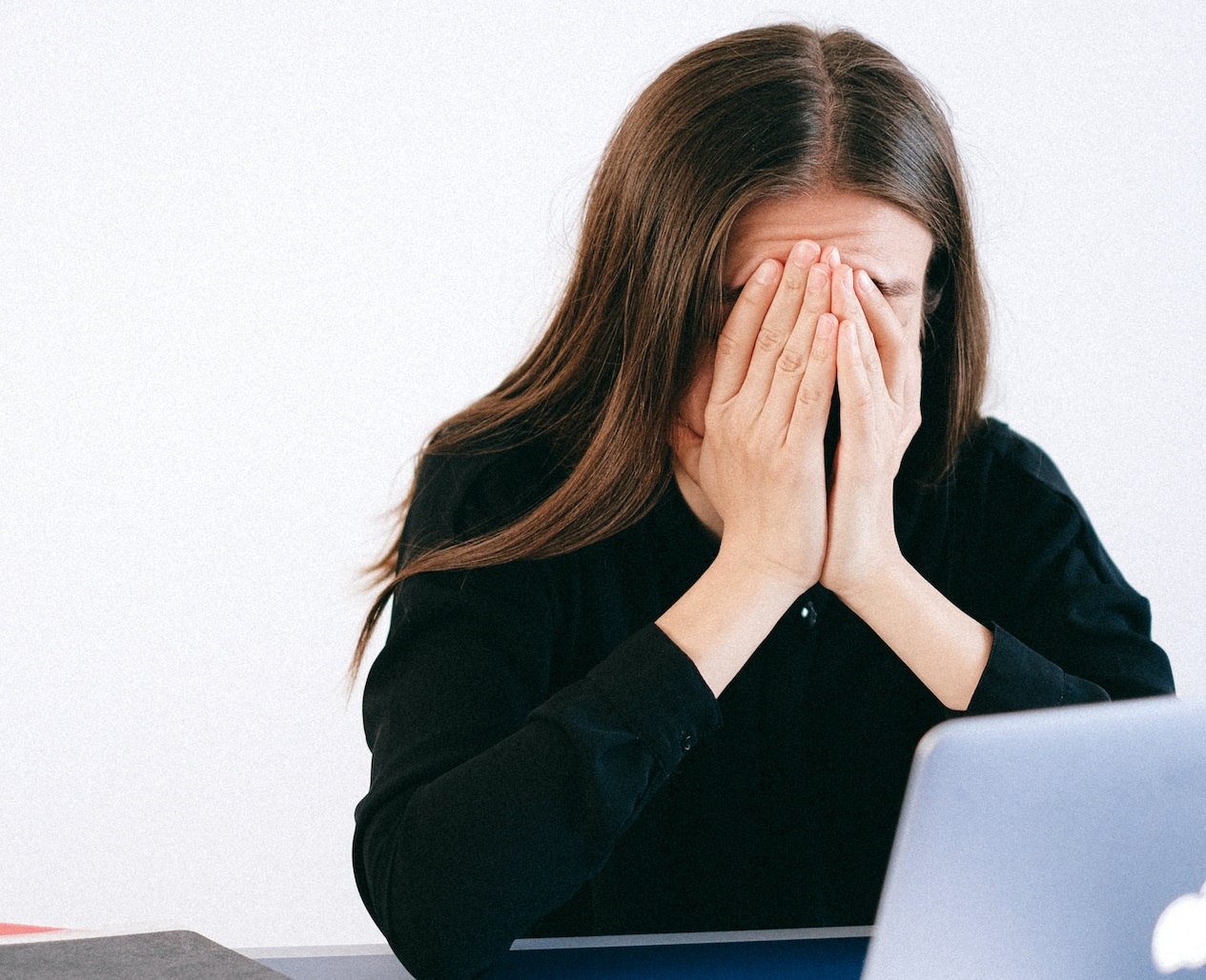 Woman is covering her face with hands and seating on the desk.