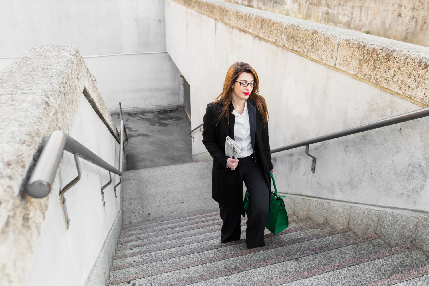 Young business woman with bag and newspaper is walking up on stairs to work.