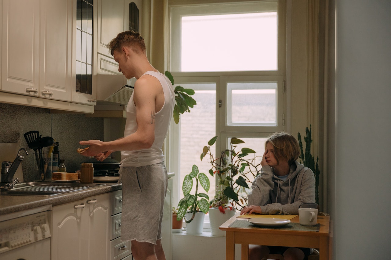 Older brother is cooking a meal to his small brother , seating on the table.
