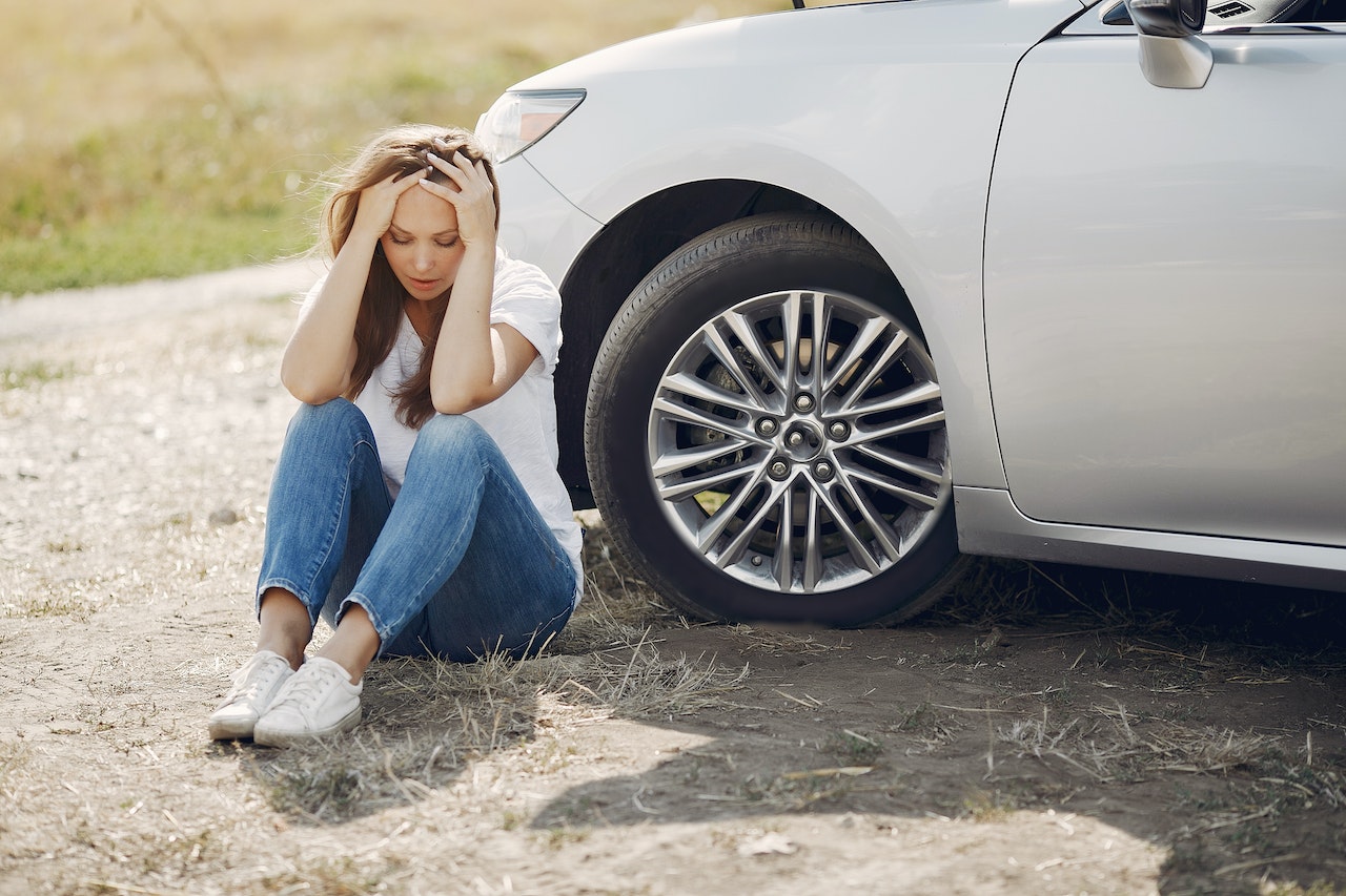 Young girl is seating on the ground next to her broken car.