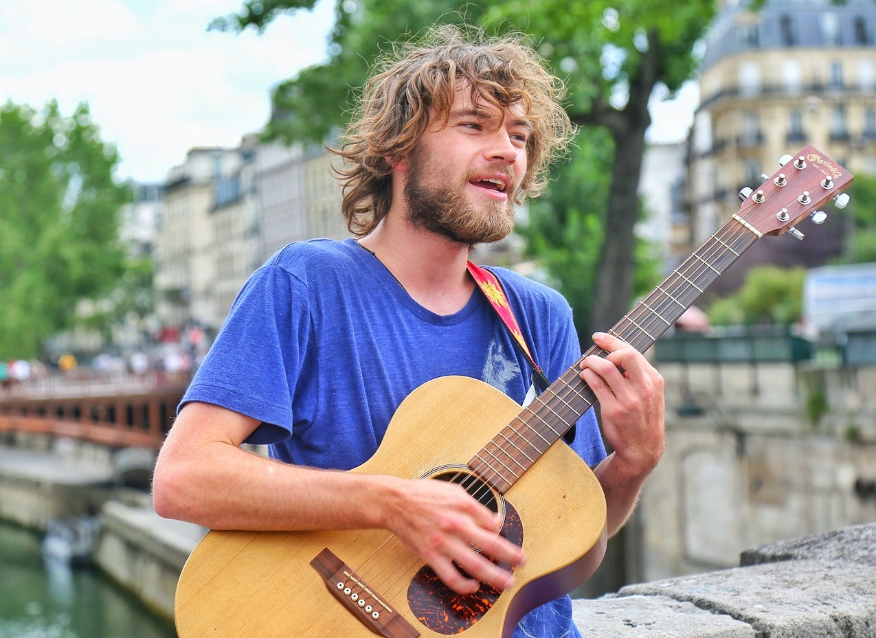 Man wearing blue shirt is playing on guitar outside on the street.