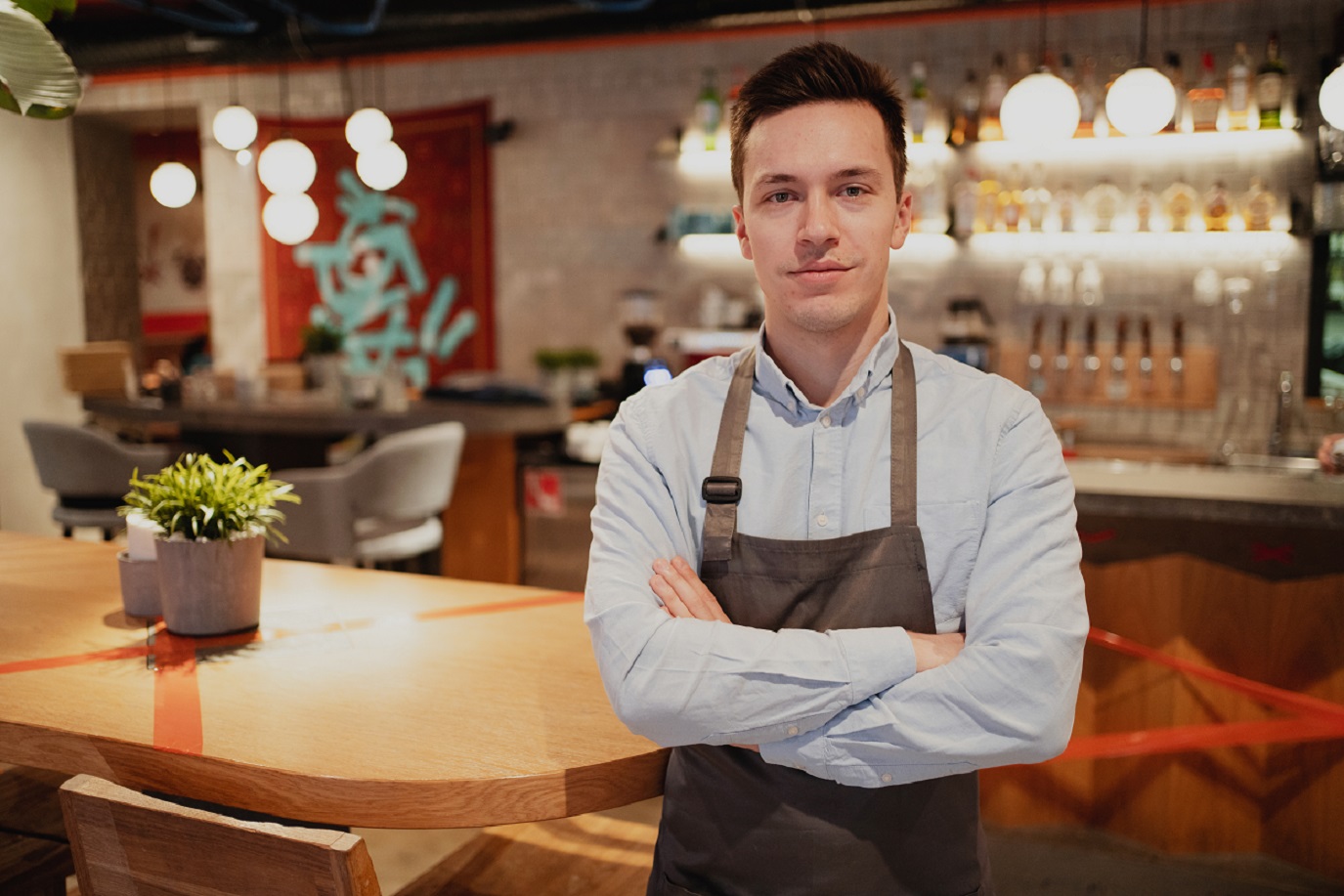 Young waiter is standing in empty restaurant and looking at camera.