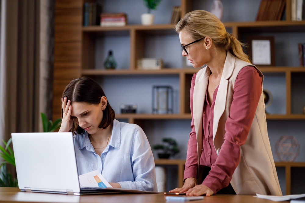Demanding female manager leader angry standing over and employee sitting at her desk watching her work