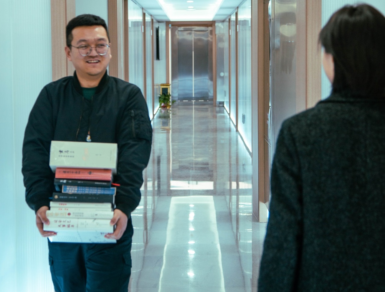 Man is holding a books and walking at hallway smiling.