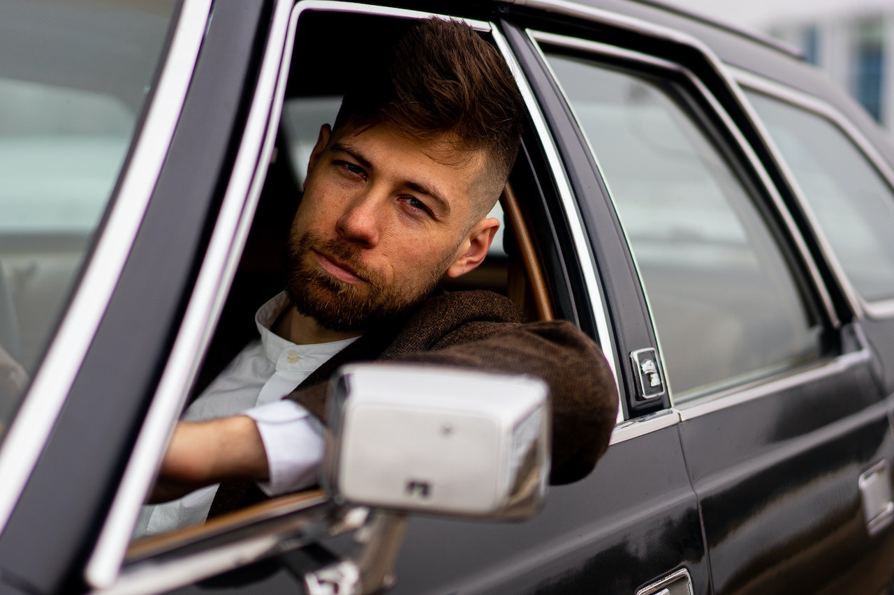 Young bearded man wearing white shirt and dark jacket is seating in black car.