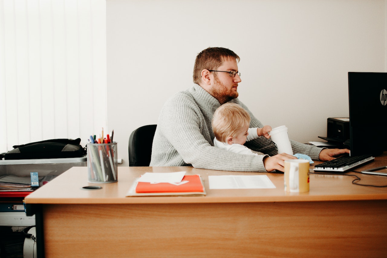 A bearded man is taking care of his son while is working at office.