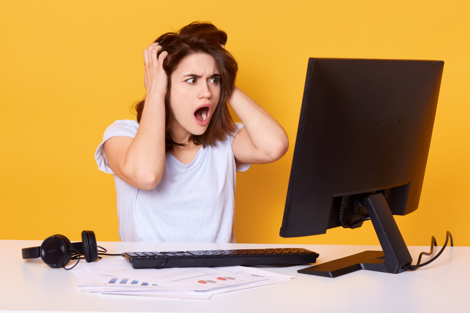 Surprised beautiful brunette girl with hands on head and open mouth is sitting in front of computer.
