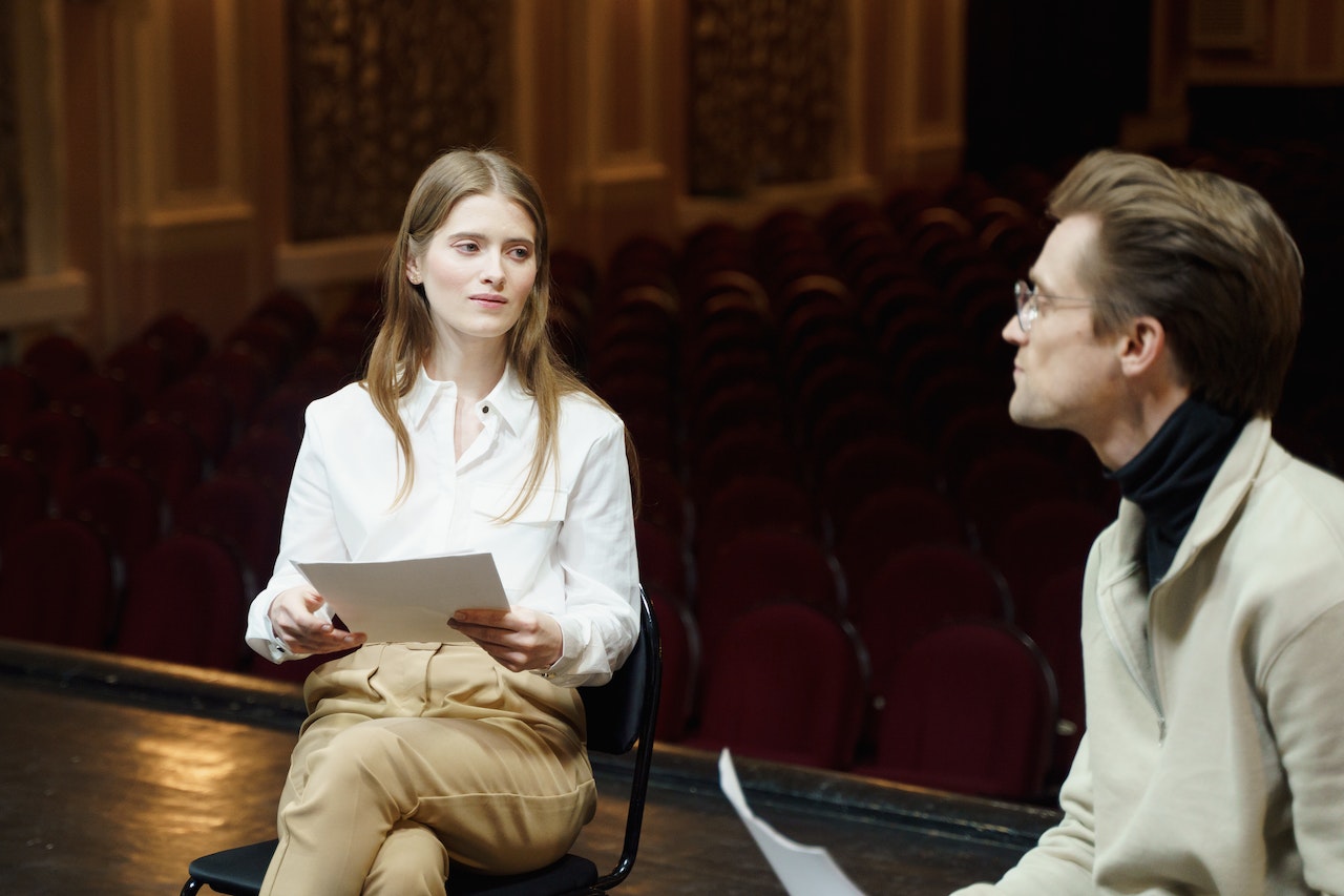 Woman is sitting on chair on theater stage and talking to other man.