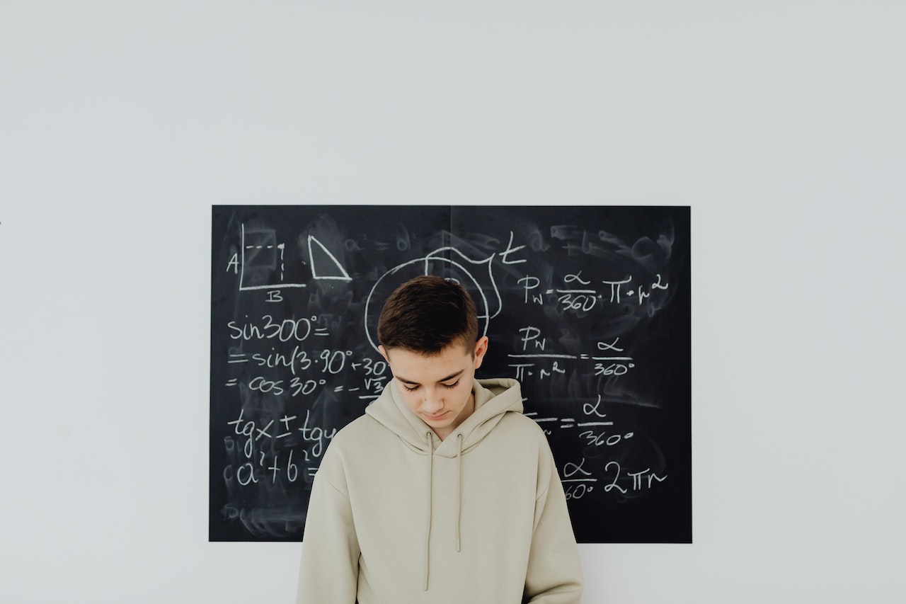 Young kid is standing sad in front of a blackboard in classroom.