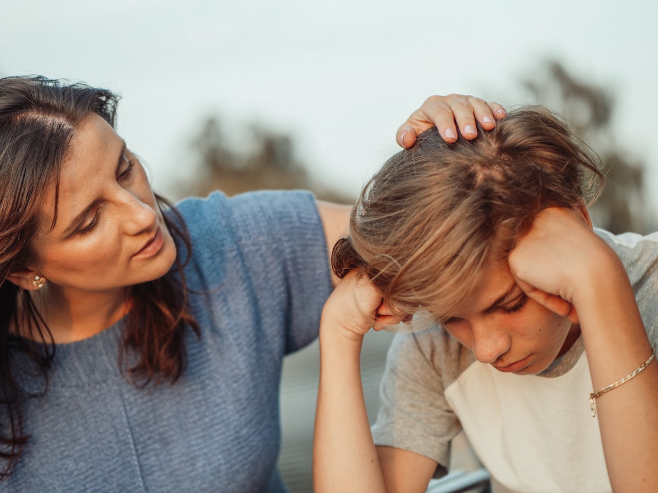 Sad kid wearing white shirt is talking with his mother outside.