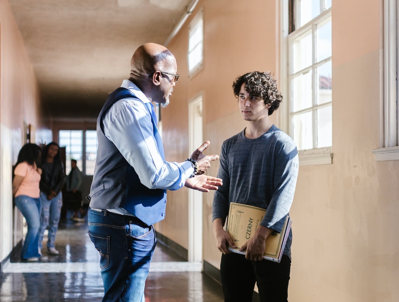 Teacher in blue shirt is talking with student in school hallway.