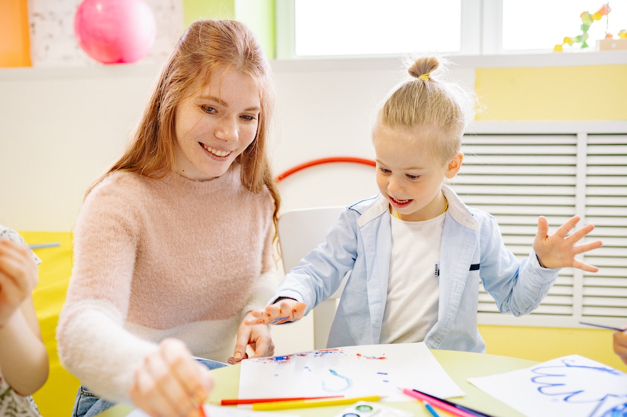 Young teacher with long hair is playing with small kid in kindergarten.