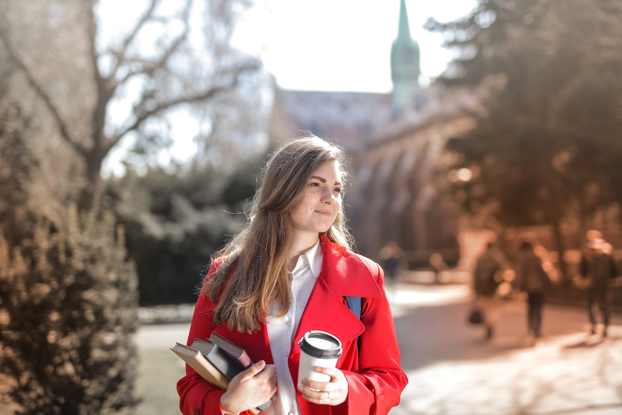 Young woman is holding books and coffee and smiling outside.
