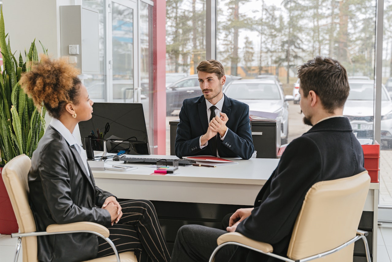 Man and woman are seating and talking with man seating on his desk.