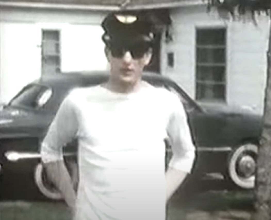 Screenshot: Buddy Holly wearing white shirt and sunglasses is standing outside - from The Real Buddy Holly Story (1985)