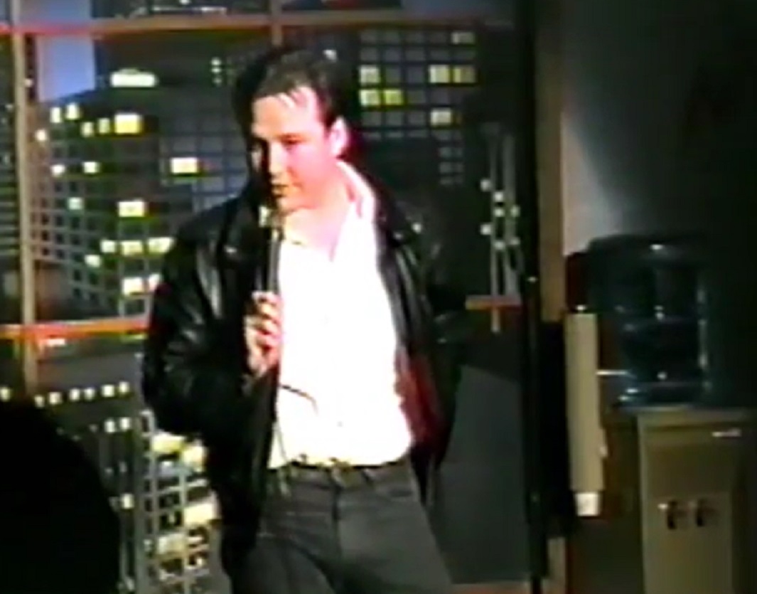 Screenshot: Bill Hicks wearing white shirt and black jacket is performing on stage - from American- The Bill Hicks Story (2009)