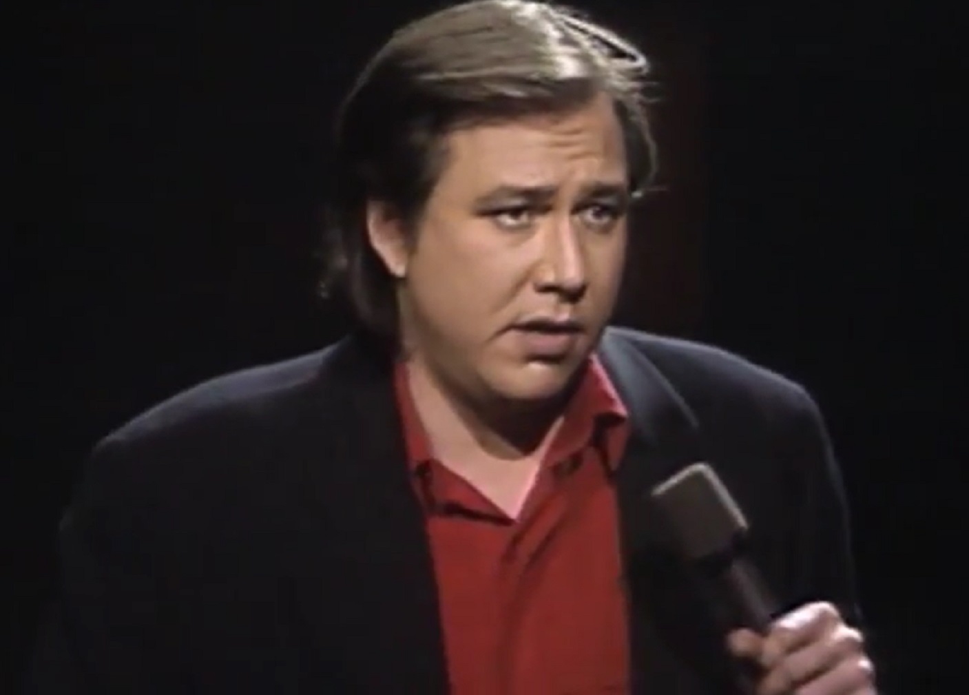 Screenshot: Bill Hicks wearing red shirt and black jacket is performing on stage - from American- The Bill Hicks Story (2009)