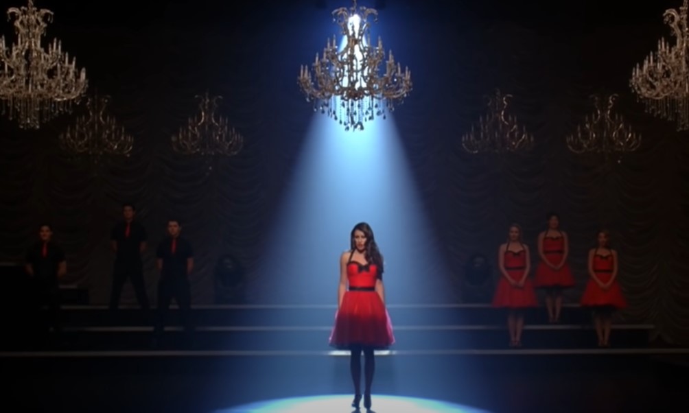 Lea Michele as Rachel Berry in red dress singing on stage with light on top of her looking