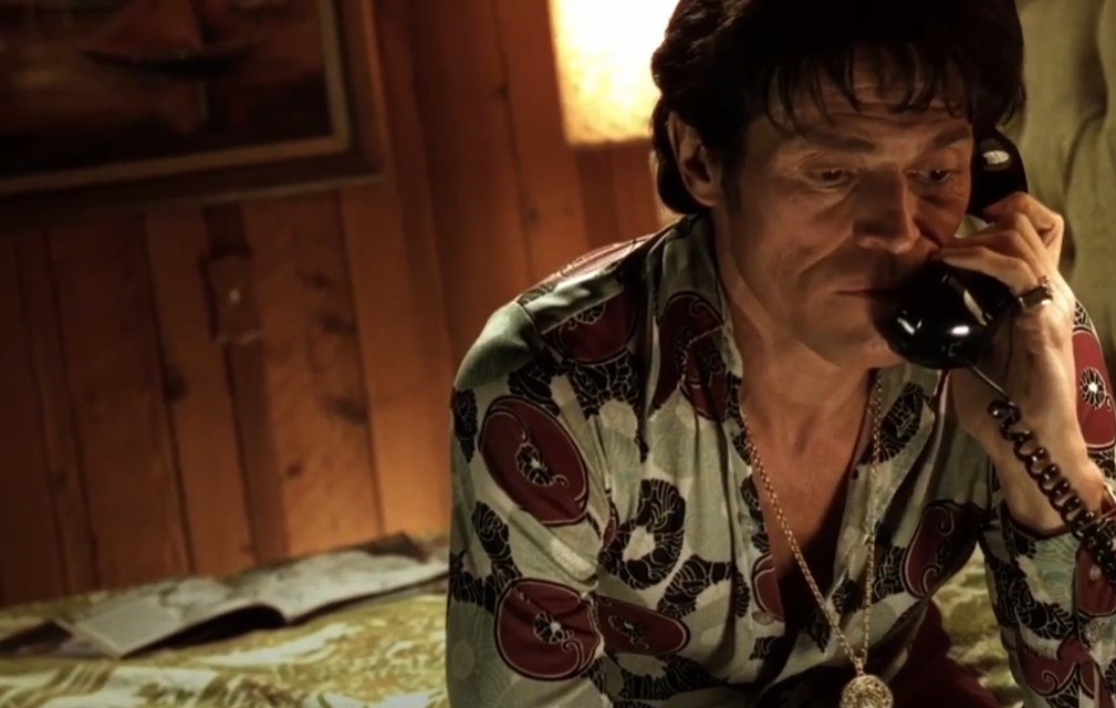 Willem Dafoe  as John Carpenter sitting on a bed